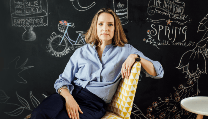 Sarah Brown, founder of Pai Skincare, seated in blue shirt against chalkboard wall with spring illustrations
