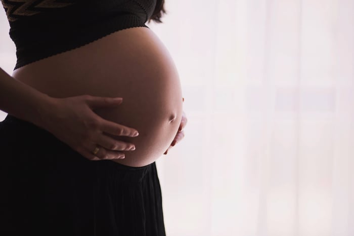 Pregnant woman in black top cradling baby bump with both hands against soft pink background