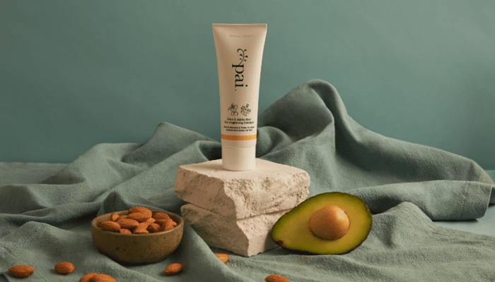 Pai Skincare Kukui & Jojoba Bead Skin Brightening Exfoliator tube displayed on textured stone with halved avocado and almonds on sage green fabric