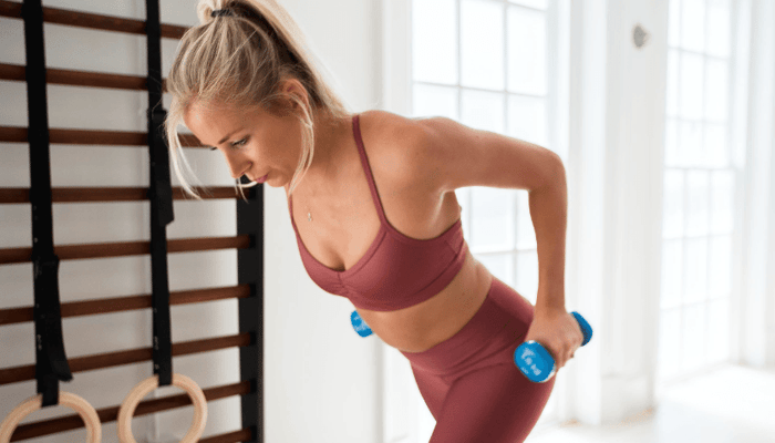 Blonde woman exercising with dumbbells in burgundy activewear in bright home gym