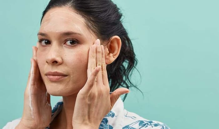 Unretouched portrait of woman applying skincare product to face against mint green background showing real skin texture