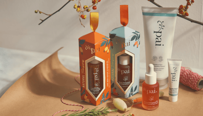 Pai Natural Treasures Christmas gift boxes with festive orange and teal packaging, displaying skincare products including oils and cleanser on warm background with seasonal branches