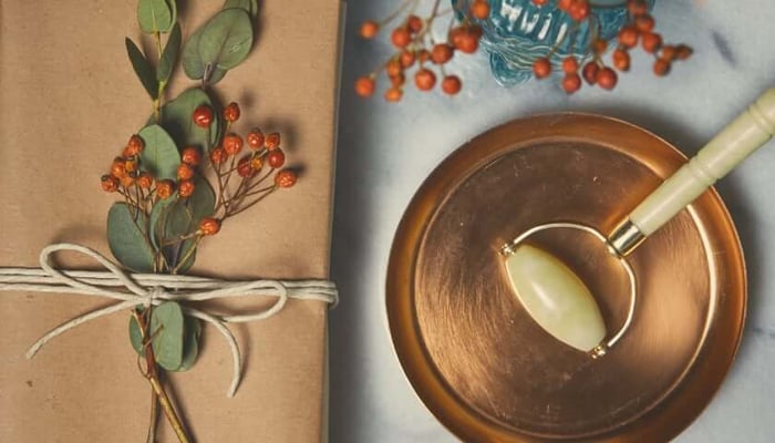 Jade facial roller with cream handle resting in wooden bowl alongside wrapped gift with orange berry sprigs