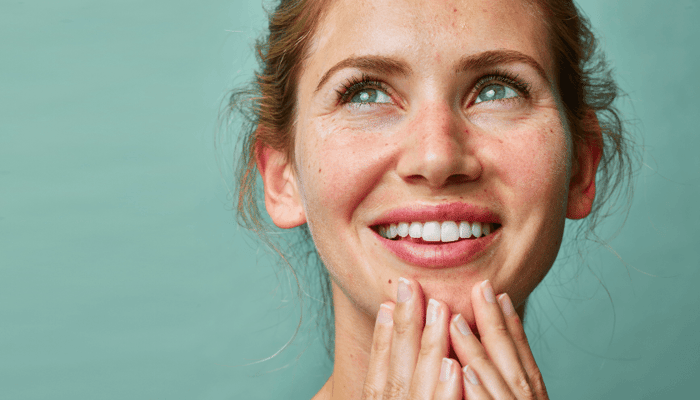 Young woman with clear, radiant skin smiling and gently touching her chin against mint green background
