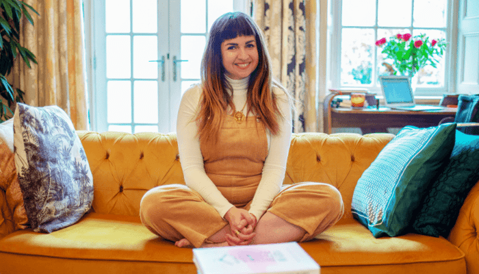 Life coach Lucy Sheridan sitting cross-legged on yellow sofa in bright living room, sharing wellness and skincare tips