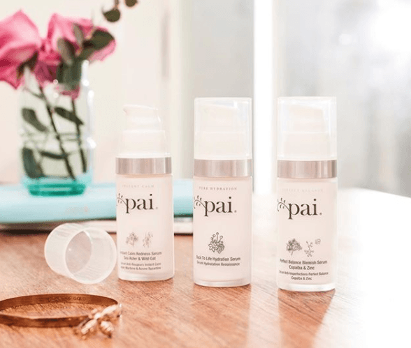 Three Pai Skincare serum bottles arranged on wooden surface with pink roses and natural light from window