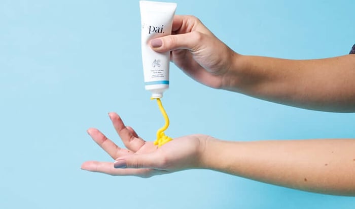 Hand squeezing golden Pai Buriti Balm from white tube onto palm against soft blue background