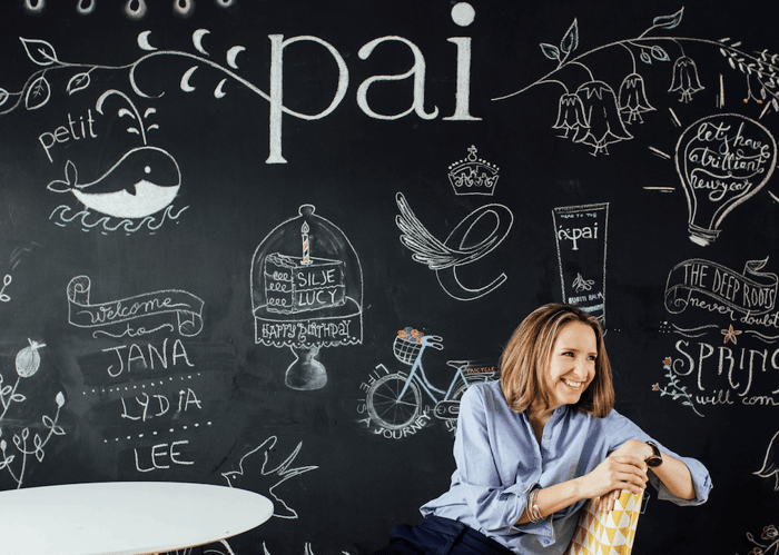 Sarah Brown, Pai Skincare founder, smiling warmly while sitting against creative chalkboard wall featuring hand-drawn Pai branding and botanical illustrations