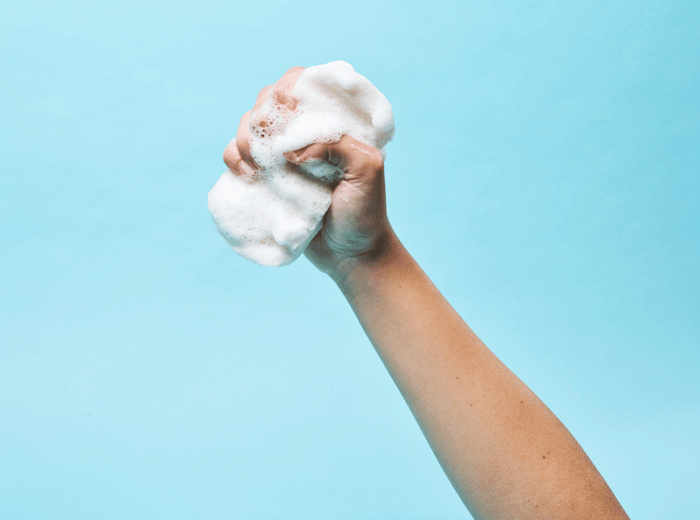 Hand holding white konjac sponge with rich foam lather against soft blue background