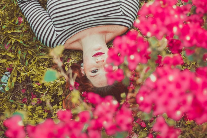 Woman lying among vibrant pink flowers in striped shirt, representing natural beauty and clean skincare lifestyle