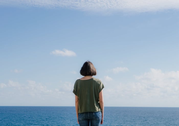 Woman with short bob haircut in olive green top looking out at calm blue ocean, symbolizing skin wellness journey