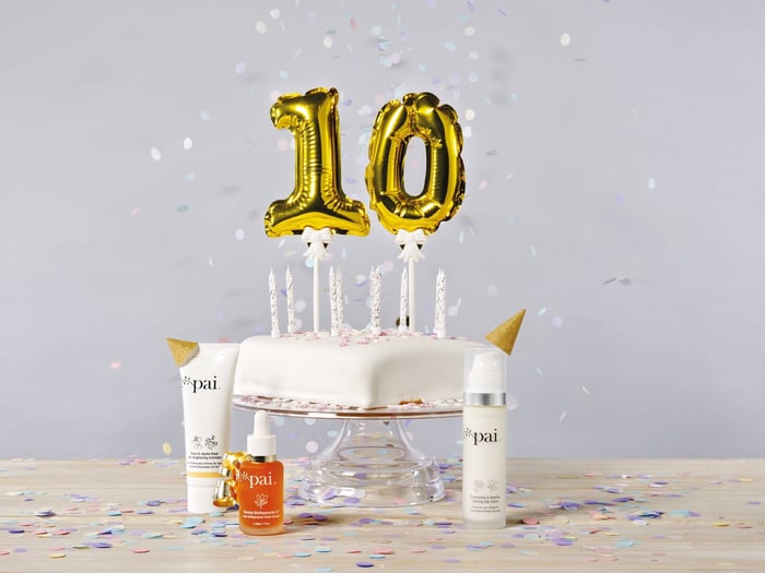 Pai Skincare 10th anniversary celebration with gold number balloons, white cake with candles, and organic skincare products surrounded by confetti