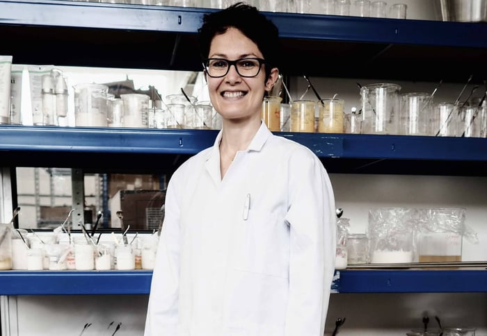 Jeanne-Marie, Pai Skincare formulator, smiling in laboratory wearing white lab coat surrounded by ingredient samples and skincare formulations