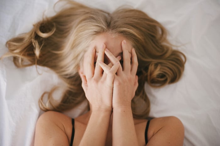 Woman lying on white bedding covering her face with both hands in embarrassment, illustrating facial blushing and sensitivity
