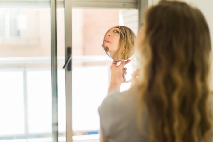 Woman examining her face in a round mirror by bright window, checking skin condition
