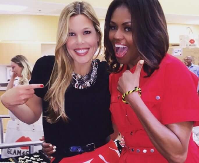 Two women smiling at beauty event - blonde woman in black dress with statement necklace and brunette woman in red dress making excited gestures