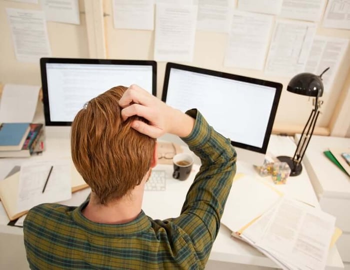 Person with hand on head showing stress at desk with computer monitors, illustrating skin condition frustration