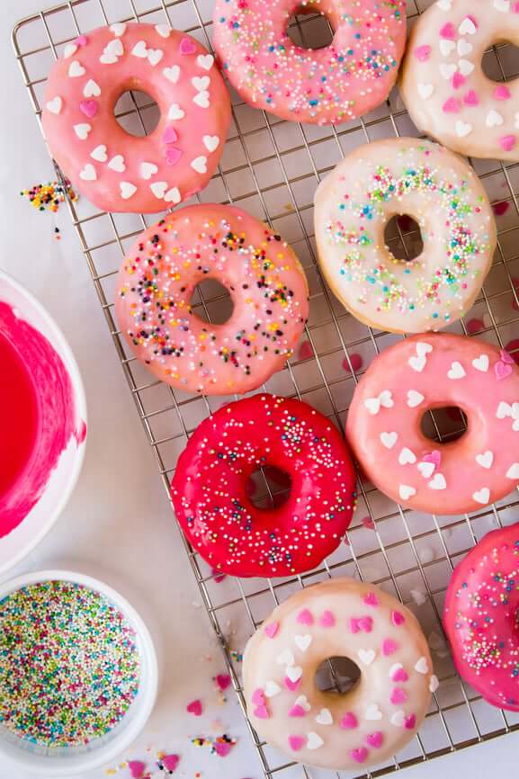 Colorful glazed donuts with sprinkles on cooling rack beside pink icing bowl - refined sugar treats