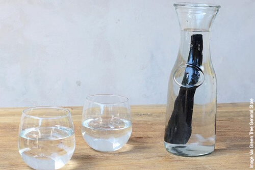 Glass carafe with activated charcoal water filter and two glasses of clear filtered water on wooden surface