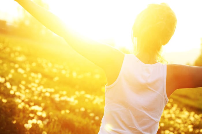 Woman in white top stretching arms toward bright golden sunlight, natural vitamin D synthesis for healthy skin