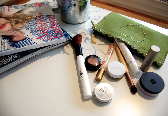 Natural makeup flatlay on white surface featuring clean beauty products including RMS compact, Pai skincare, and green studded makeup bag