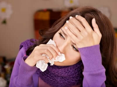 Woman with seasonal allergies holding tissue to nose while touching forehead, wearing purple sweater