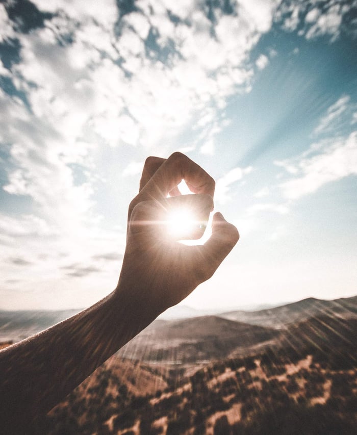 Hand reaching toward bright sunlight against blue sky and mountainous landscape, illustrating sun exposure and photosensitive skin reactions