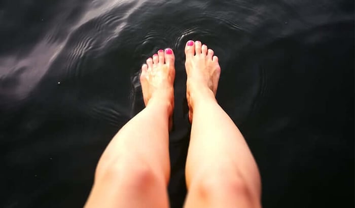 Bare legs and feet with pink nail polish dangling in dark water, showing self-care moment