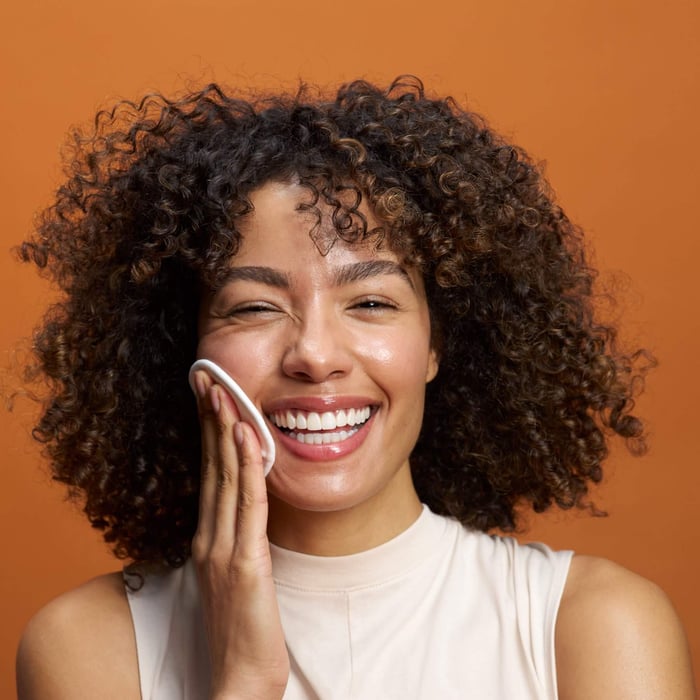 Smiling woman with curly hair applying Pai Bright Now PHA Exfoliating Toner with cotton pad against warm orange background