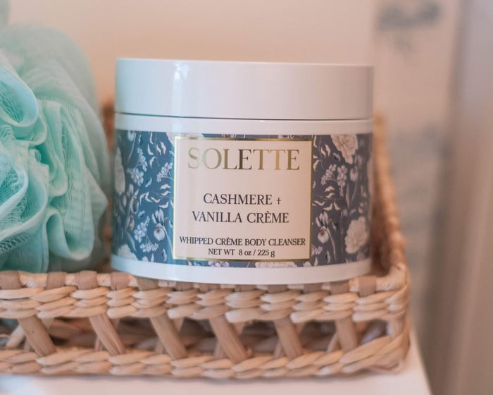 jar of cashmere and vanilla creme body wash on a counter with a bath sponge and a rattan tray