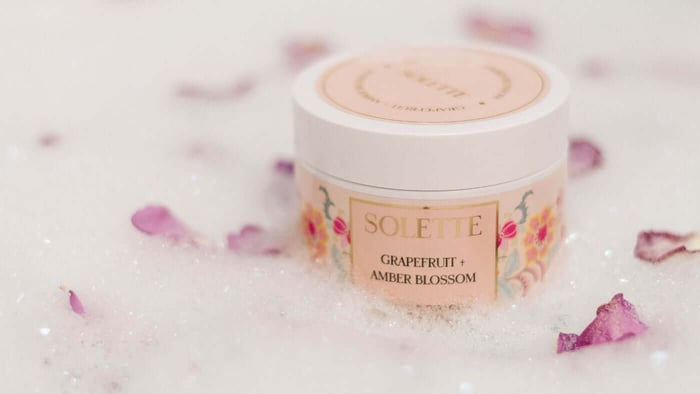 a jar of grapefruit and amber blossom sugar scrub floating in a bubble bath with flowers for a spa night at honme