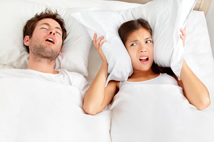 Woman covering her ears with a pillow while her partner sleeps next to her, snoring with his mouth open. 