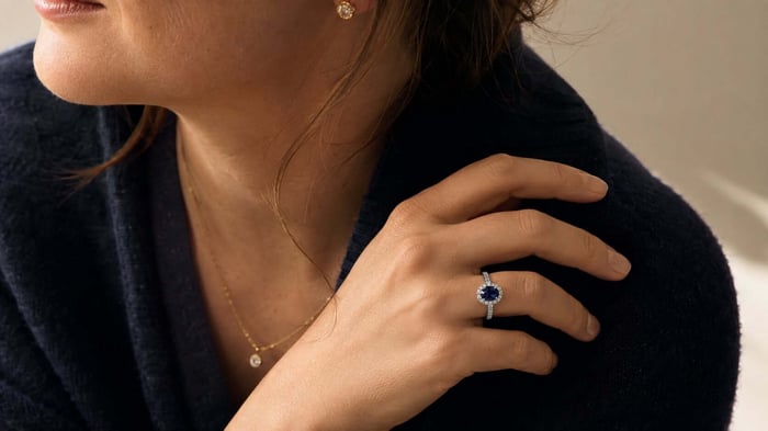 A blue sapphire and diamond ring worn with simple yellow-gold diamond jewellery and a restrained dark outfit.