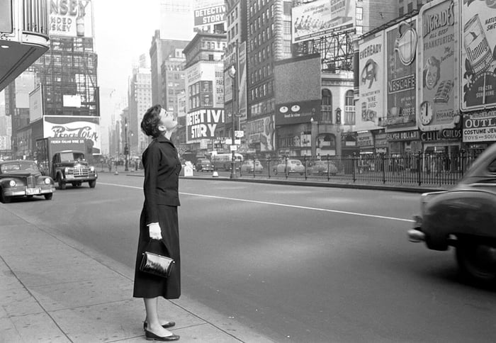Audrey Hepburn stands in Times Square in 1951. She looks up at the billboards as cars pass by. 