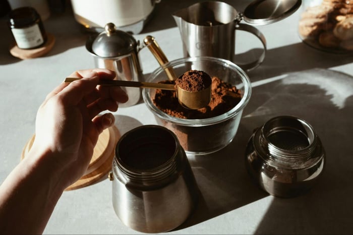 Hand scooping ground coffee into the moka pot under warm morning light