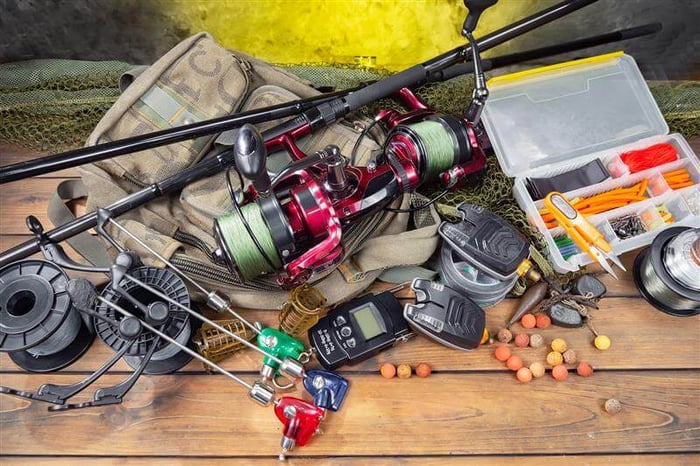 Assortment of fishing tackle, reels, bite alarms, and bait organized on a wooden surface.