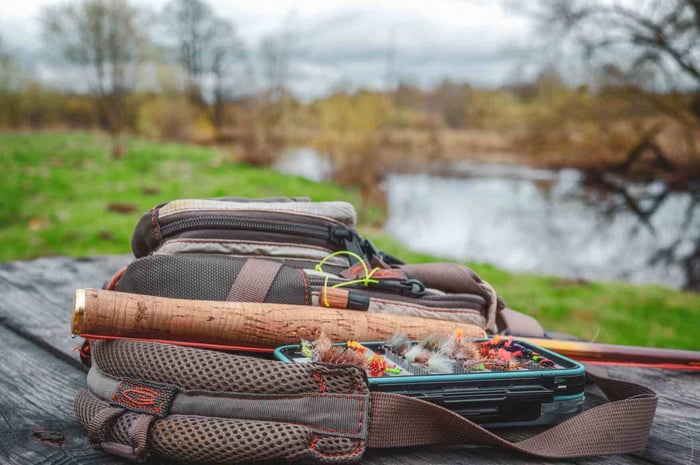 Fly fishing gear with a tackle box and rod placed on a wooden table beside a riverbank.