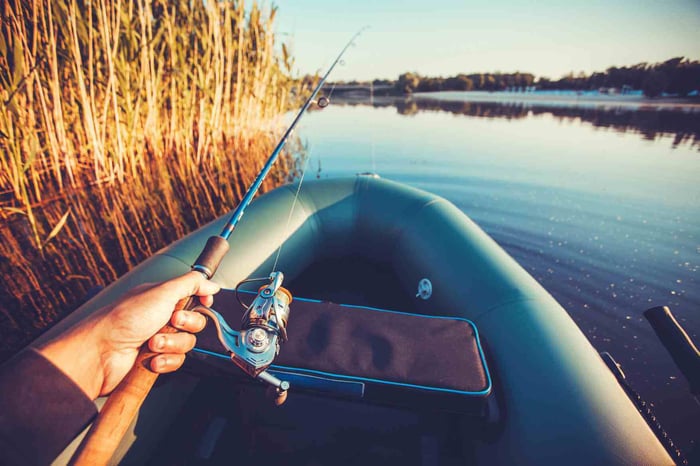 Angler holding a fishing rod on an inflatable boat near a calm lake shoreline at sunrise.