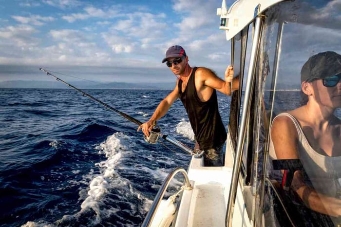 Man deep sea fishing from a boat on a sunny day, holding a fishing rod while another person steers.