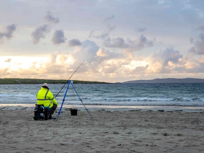 Person in a high-visibility jacket beach fishing at sunset with a rod on a tripod.