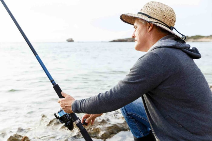 Man in a straw hat fishing from a rocky shoreline, holding a rod and looking at the water.