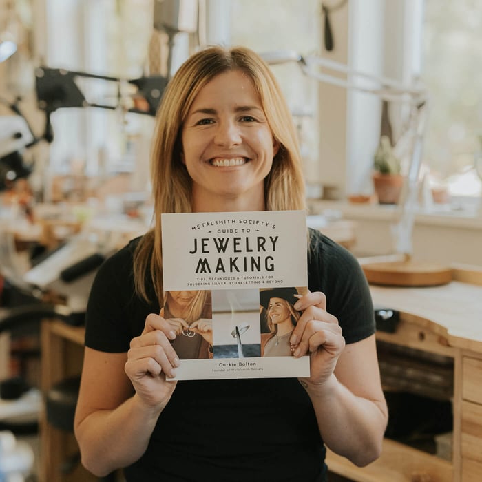 Corkie Bolton, author of Metalsmith Society's Guide to Jewelry Making, holding the book and smiling, showcasing the cover and sharing her passion for jewelry craftsmanship.