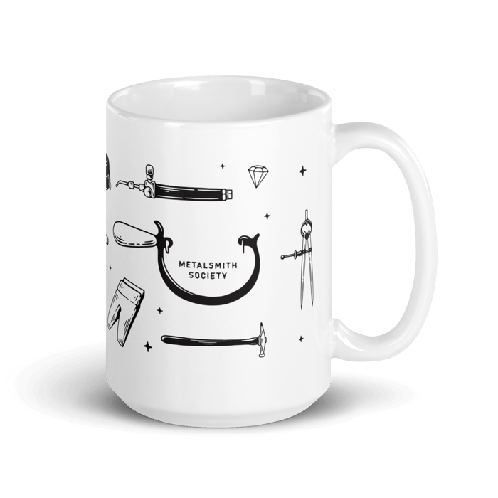 White Metalsmith Society Mug with jewelry making tools including a saw frame, calipers, a bench pin, a torch head and a jeweler's hammer. The mug is on a white background.