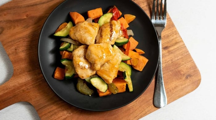 Balanced prepared meal portions with grilled chicken pieces served over zucchini, sweet potatoes, and peppers on a black plate.