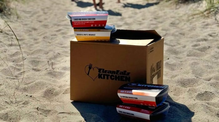 Weight loss prepared meal delivery showing Clean Eatz meal containers stacked beside a branded box on a sandy beach.