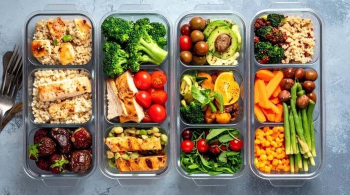 Portion-controlled meals lined up in glass containers with grilled chicken, quinoa, broccoli, avocado, carrots, and roasted vegetables.