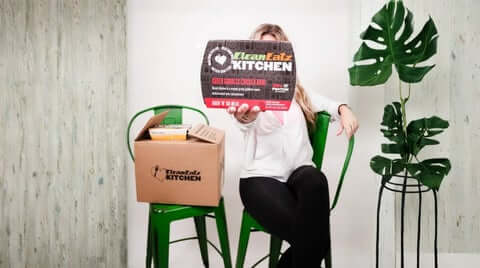 Best premade meal delivery showing a person holding a Clean Eatz Kitchen packaged meal in front of a home setup with a branded delivery box.