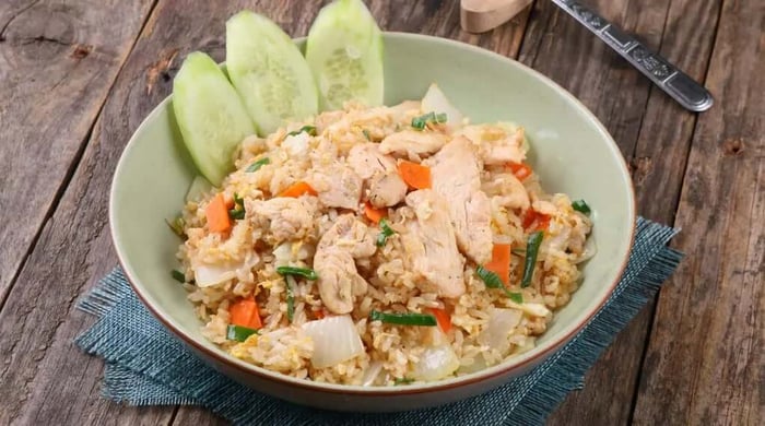 Bowl of chicken stir-fry with sugar snap peas, carrots, onions, and egg tossed with fried rice and served with fresh cucumber slices on a rustic wooden table.