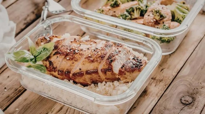 Sliced grilled chicken breast over white rice with fresh basil in a healthy meal delivery container on a wooden table.