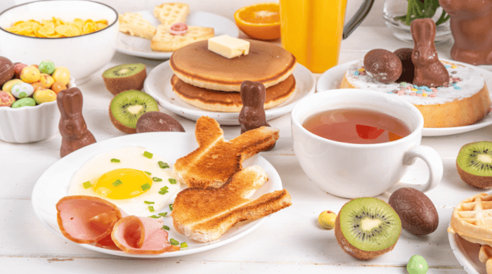 Healthy breakfast table spread with fried eggs, ham, bunny-shaped toast, pancakes, waffles, kiwi, orange juice, and chocolate Easter bunnies.